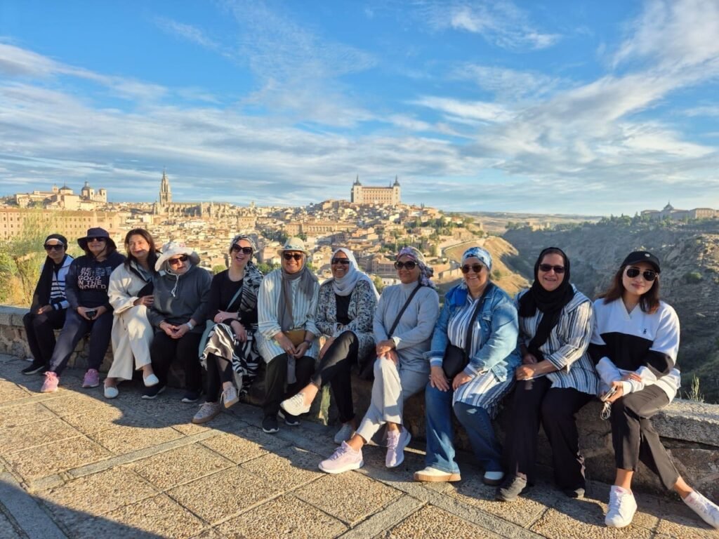 A group of female travelers wearing modest clothing and hijabs posing together on a stone wall overlooking the historic skyline of Toledo, Spain.
